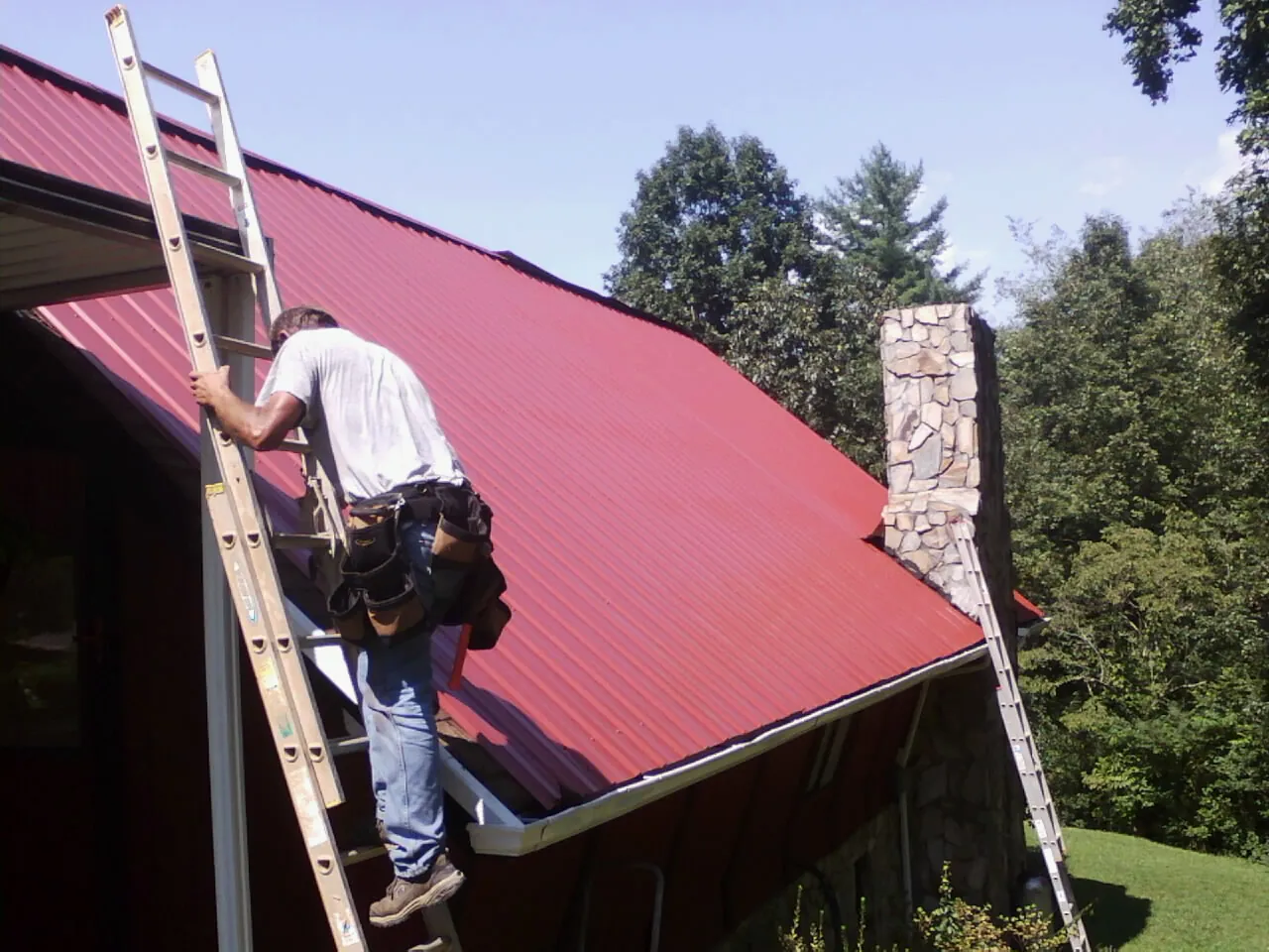 Roofer on a ladder beside a red metal roof and stone chimney — Soffit & Fascia Repair by Roofing Brothers in Pleasantville